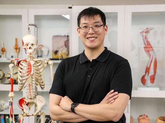 Photo of an Asian male in a black shirt standing in an anatomy lab with a model skeleton to his right.