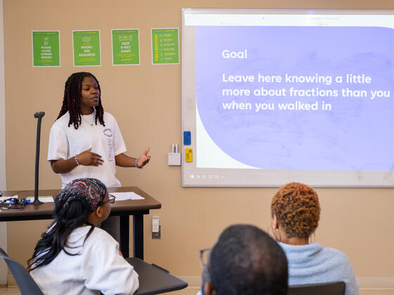 Photo of a Black student in front of a projector screen speaking to a classroom of people.