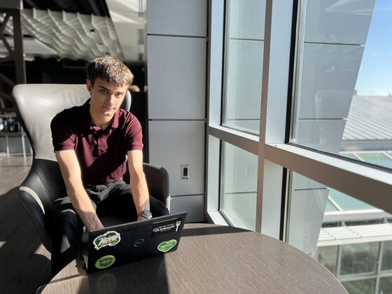 A mal student wearing a black shirt and pants sits by a window with his hands on a laptop.