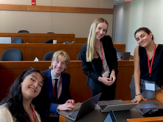 Photo of three female and one male wearing suits and lanyards gathered around a laptop while smiling.