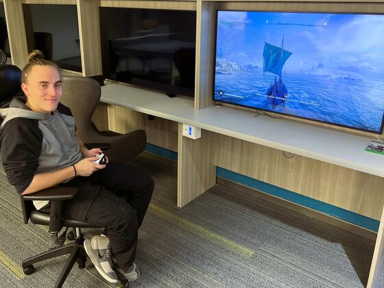 A male student holds a video game controller and smiles at the camera next to a large TV screen with a video game playing on it.