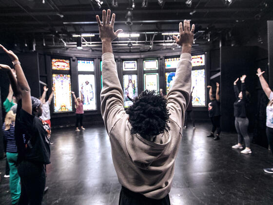 Students stand in a circle with their arms raised. The room has a panel of stained glass windows behind them.