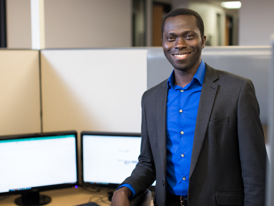 Data Analytics M.S. student Michael Eniolade wears a suit and stands next to a computer screen on a desk.