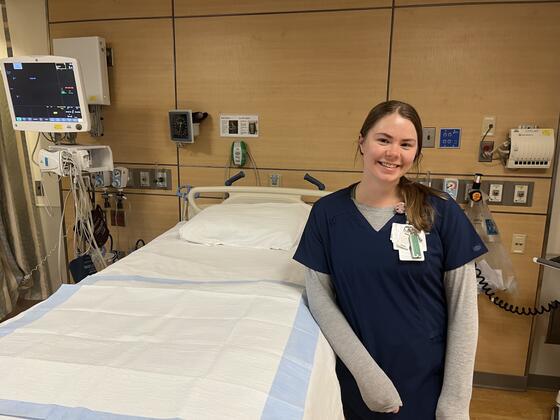 A young woman in nursing scrubs stands next to an empty hospital bed.