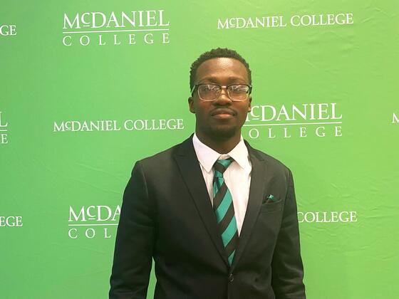 A man wears a suit in front of a green backdrop with the McDaniel College logo on it.