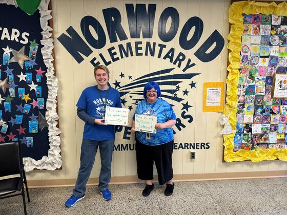 A male student stands next to a female teacher. The student holds a sign pointing to the teacher that says "this was my third grade teacher" and the teacher holds a sign that reads "This is my McDaniel intern"