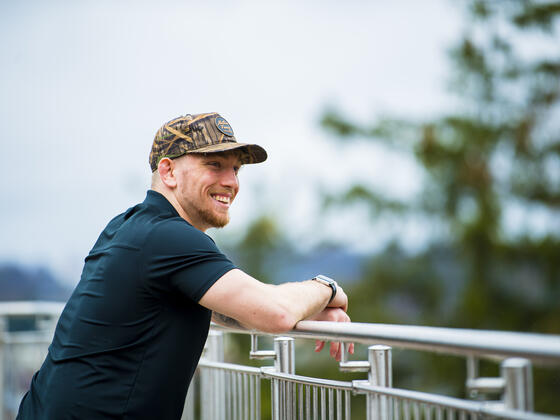A student wearing a hat leans on a railing looking off camera while smiling.