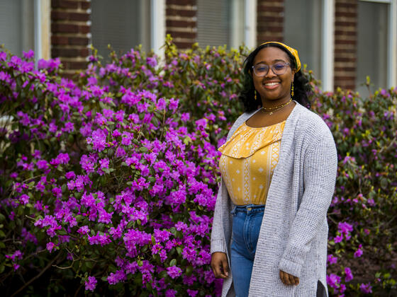A student stands in front of a purple flowering bush outside while wearing a yellow shirt and yellow bandana.