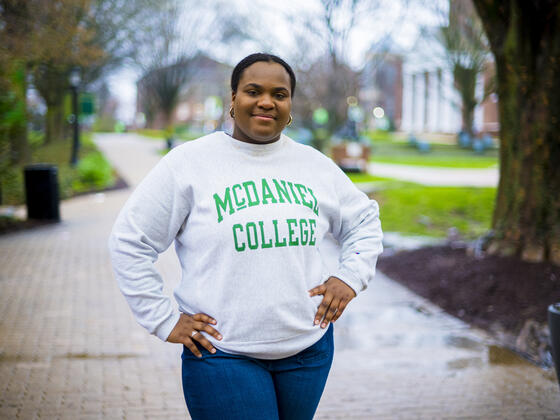 A student wearing a McDaniel College sweatshirt stands outside with her hands on her hips.