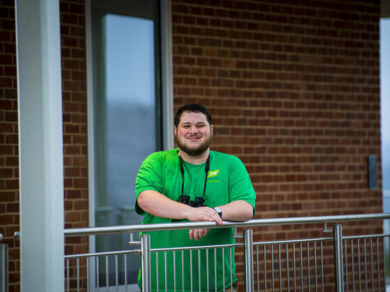 A student leans forward on a railing with a pair of binoculars around his neck.