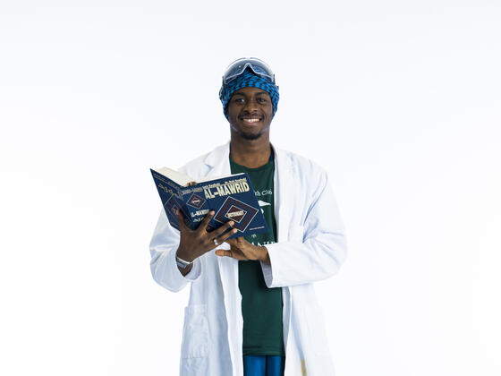 A student in a lab coat hold an Arabic book open while smiling at camera in front of a white background.