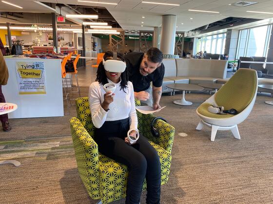 A student in black pants and a white shirt sits in a green chair while wearing a VR headset over her eyes. A middle aged male leans over her shoulder while speaking to her.