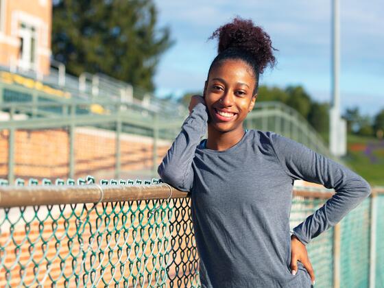 A female student in a grey athletic shirt leans her elbow on a chain link fence in front of bleachers.