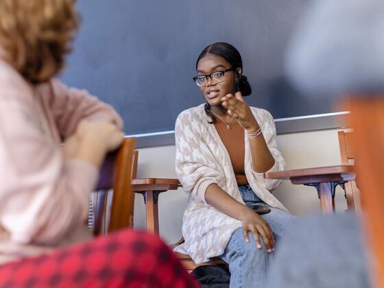 A female student in a grey cardigan seated in class speaks with one hand held up.