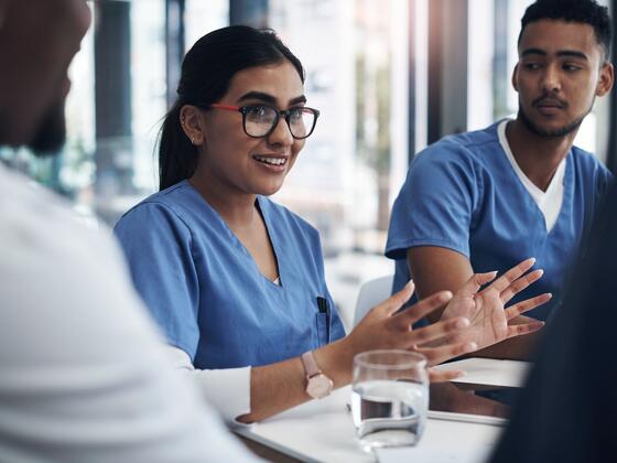 A woman in blue scrubs wearing glasses holds her hands out while speaking at a table with other medical professionals.