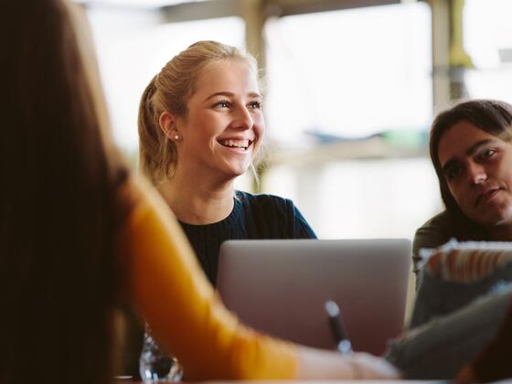 A female student sits in a classroom with a laptop in front of her, with two other students on either side of her.
