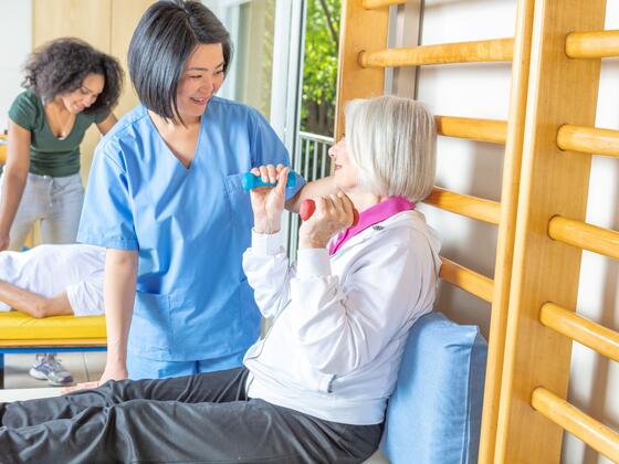 A woman in blue medical scrubs stands besides an elderly woman who is seated and holding up small dumbbells in her hands.