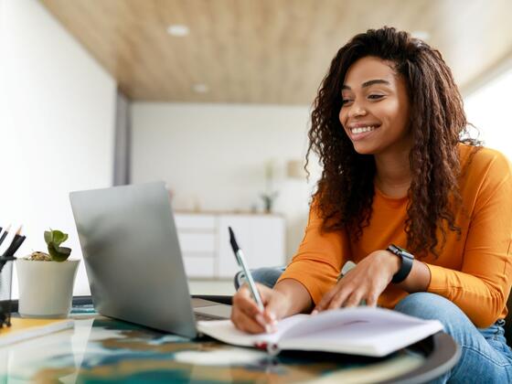A woman in an oragne long-sleeved shirt smiles at a laptop screen while taking notes in a notebook.