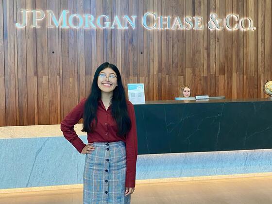 A Hispanic woman in a grey skirt and red blouse smiles in front of a counter, with a sign on the wall behind her that sayd "J.P. Morgan Chase and Co."