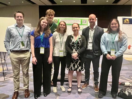 five students and two professors standing by posters at a conference wearing business causal clothes.