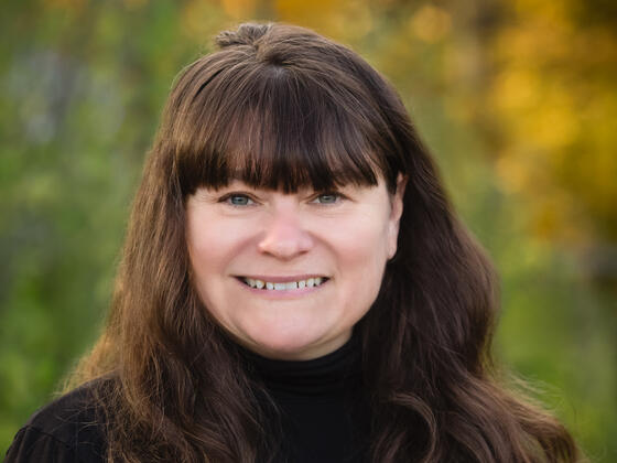 Pamela Mesta, a woman with long brown hair and bangs, wears a black turtleneck and smiles in front of an outdoor background.
