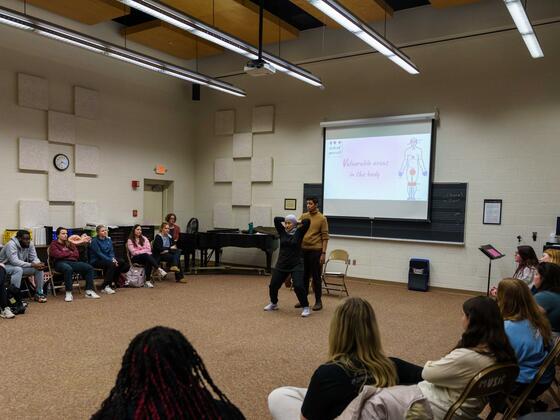 A class of students sit in a semi-circle around two self-defense instructors and a powerpoint screen showing an images labeled "vulnerable areas of the body."