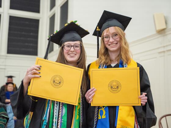 2 students in graduation regalia pose with gold award envelopes