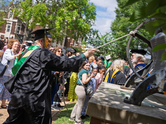 Student in graduation regalia pulls on string to ring large bell with a crowd in the background