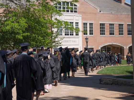 A line of graduates in regalia walk across McDaniel College's campus in front of Merritt Hall.
