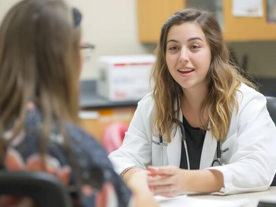 A woman in a white doctor's coat sits across a table from a patient.
