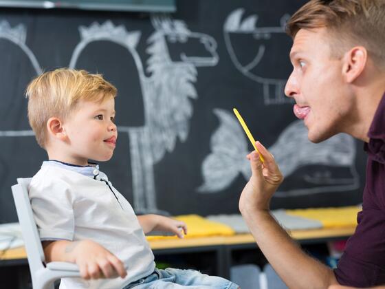 A man sits across from a little boy, holding his own tongue to his top lip, while the boy practices the same mouth shape.