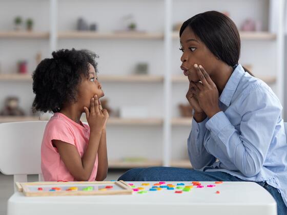 A young woman sits next to a small child at a table. They both hold their hands up to their face while shaping a sound with their mouths.