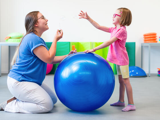 Occupational therapist kneels on the ground with a large blue ball and a young girl in pink