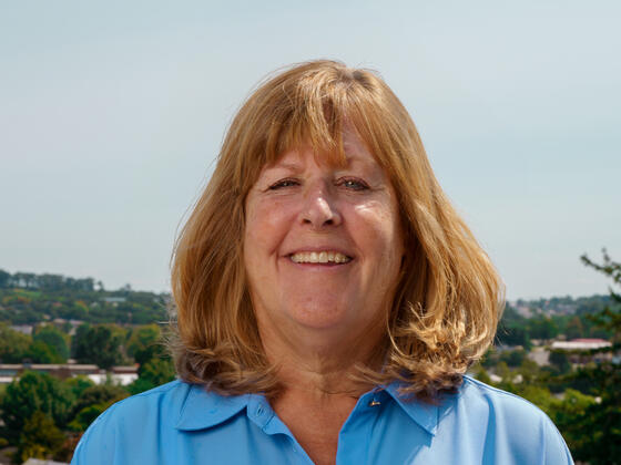 A woman with blonde hair and a blue shirt smiles in front of a scenic view.