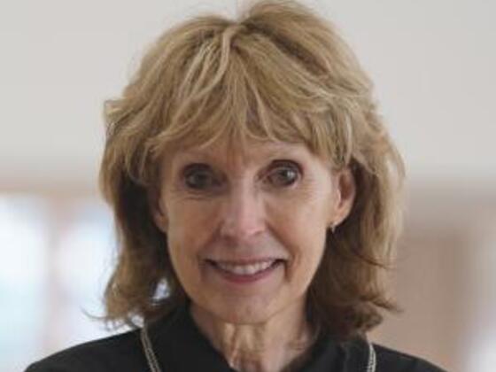 A woman with blonde hair and a black shirt smiles in front of a gray background.