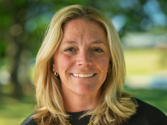 A woman with blonde hair and a black shirt smiles in front of a blurry outdoor background.