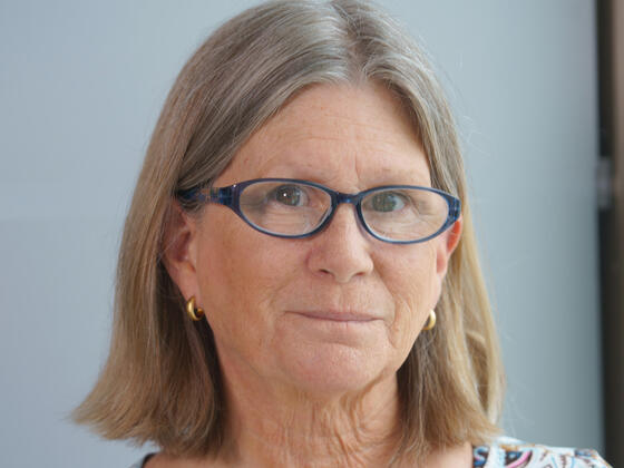 A woman with short, gray hair and glasses smiles in front of a gray background.