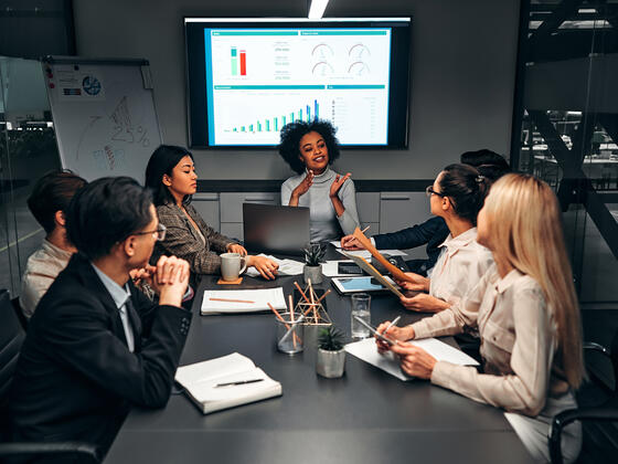 A group of diverse professionals sits around a conference room table in front of a screen of data charts.