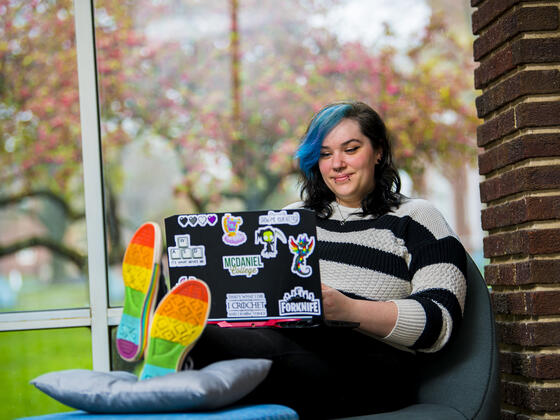 A student sits in a chair with a laptop and rainbow shoes