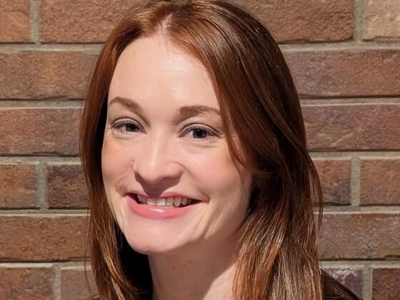 A red-haired woman smiles in a black shirt in front of a brick wall.