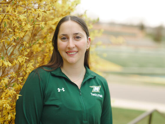 Kate Principio wears a McDaniel track and field jacket and stands in front of a yellow flowering tree.