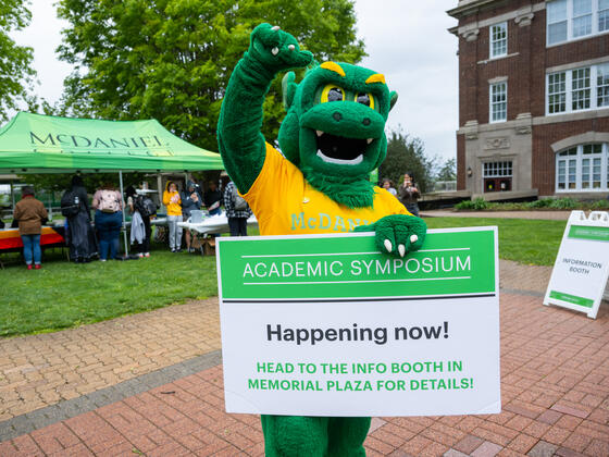 Green Terror mascot holding a sign for the Academic Symposium that reads "Happening Now! Head to the info booth in Memorial Plaza for details!"