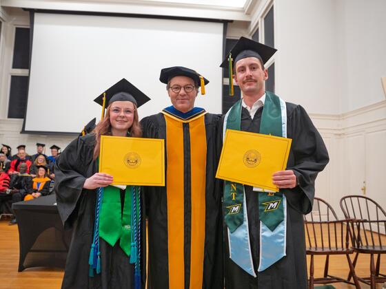 2 students in graduate regalia holding awards stand with the provost in graduation regalia with people seated in the background