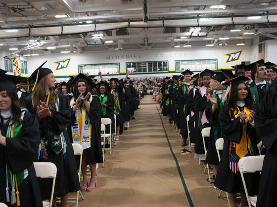 students in graduation regalia stand in rows with an aisle down the middle