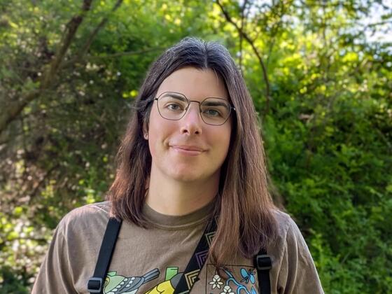 person with brown hair stands in front of trees