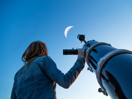 A woman holds a telescope and looks at the moon in the sky.