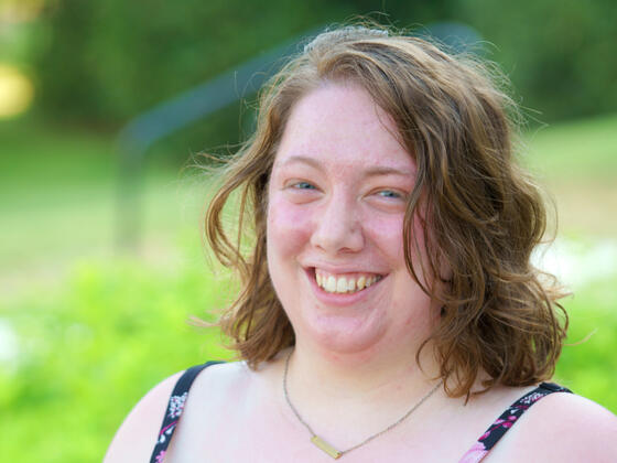 A young woman with short, brown hair wears a floral shirt and smiles in front of an outdoor background.