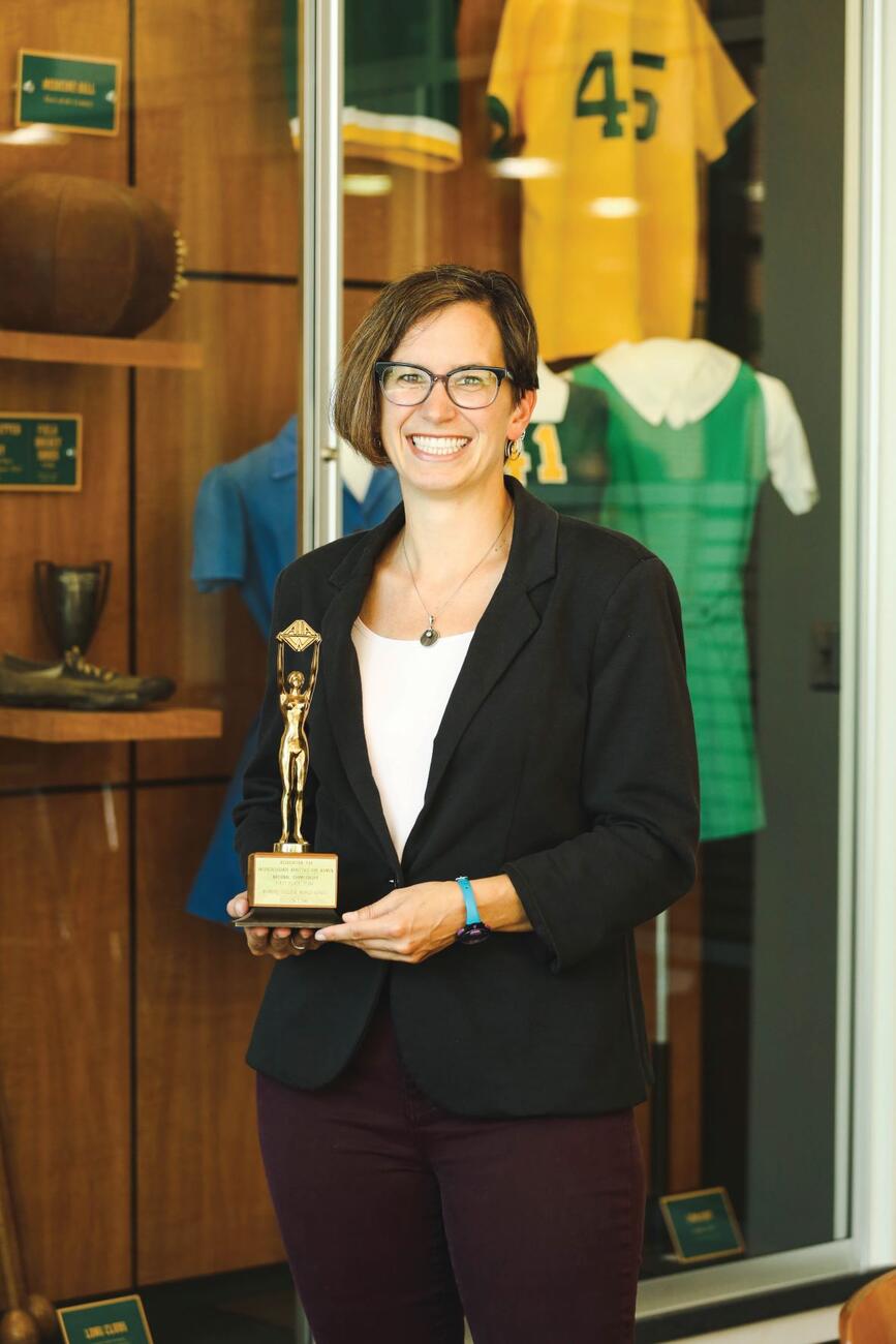 Photo of professor Diane Williams holding a trophy in front of a display of vintage McDaniel sports uniforms.