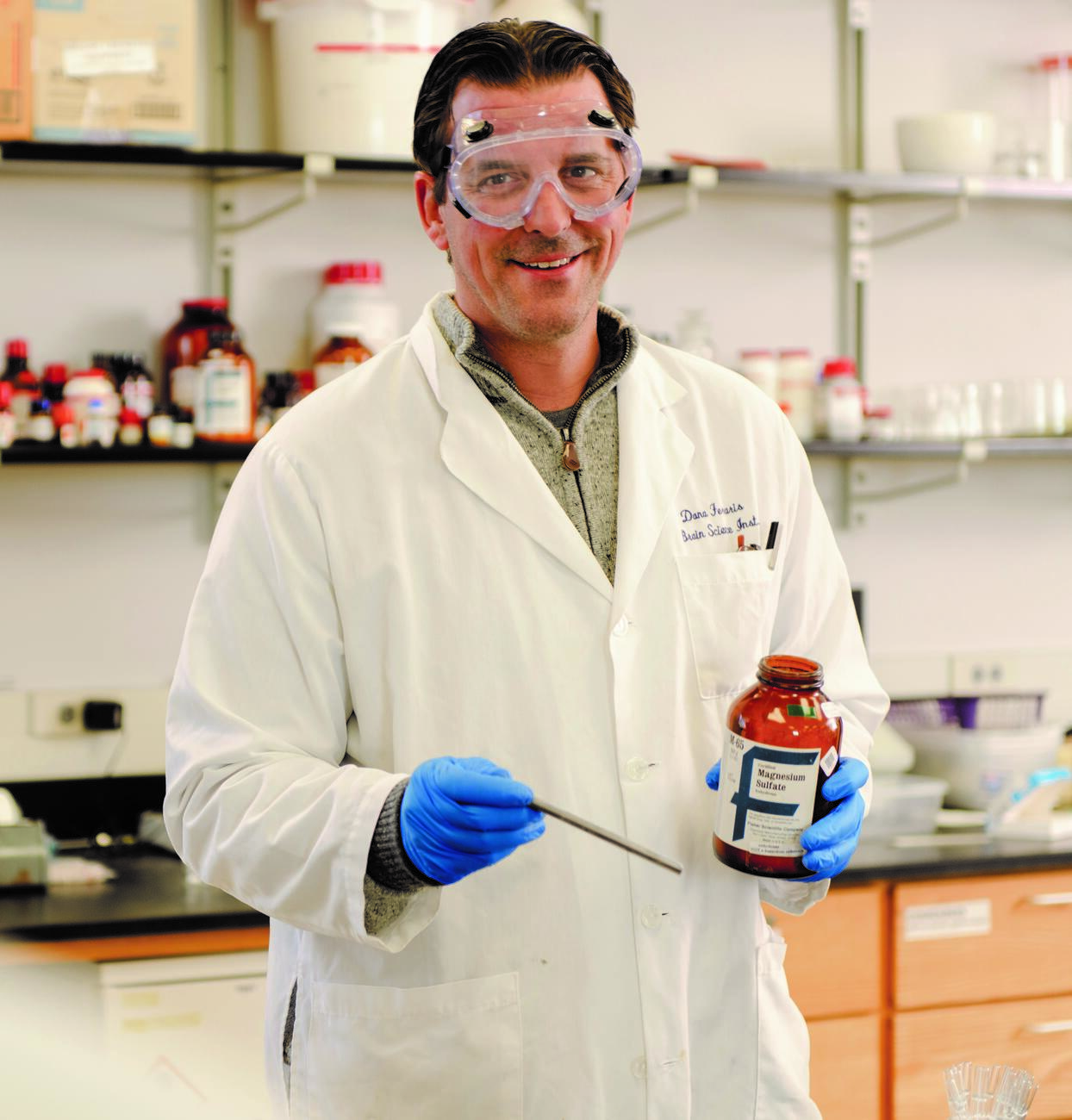 Photo of professor Dana Ferraris in a lab coat and gloves holding a bottle in a lab.