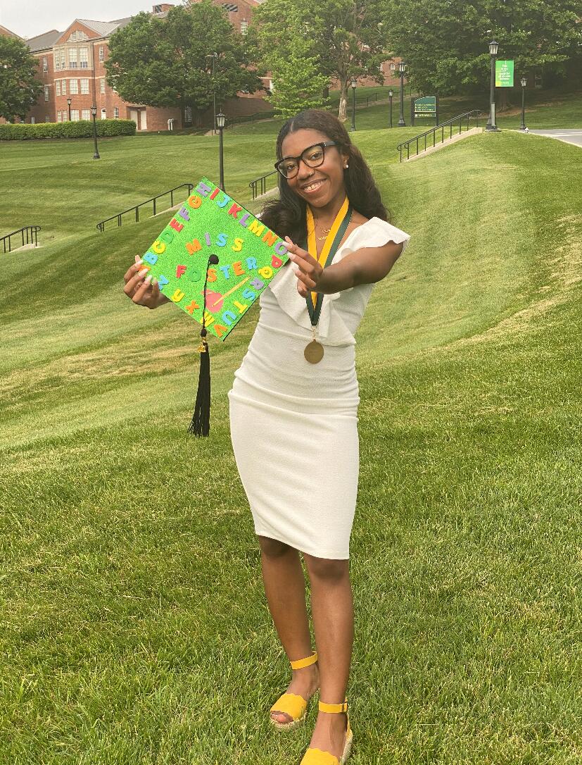 Alumna Amara Foster holding out her graduation cap, which says "Miss Foster."